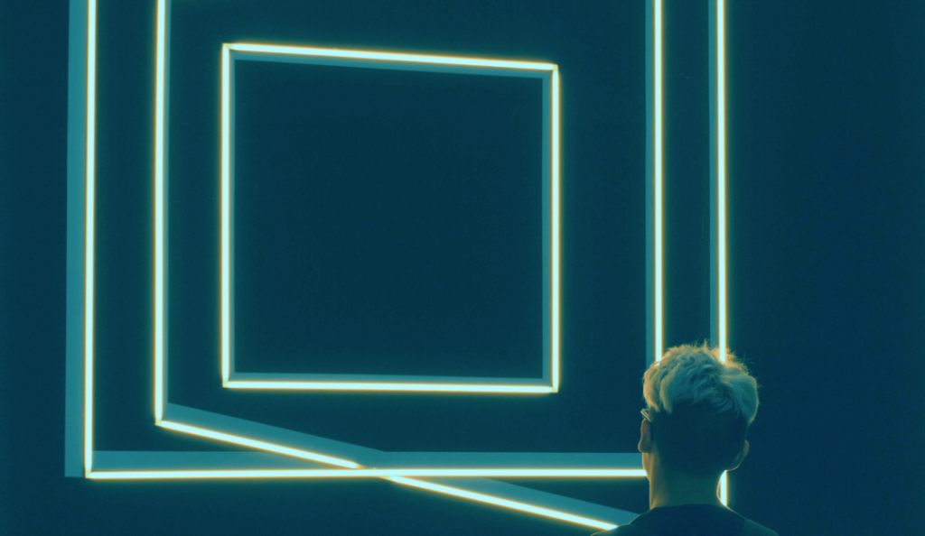 Imagem ilustrativa sobre a postagem de Design de Futuros. Na imagem, uma pessoa de cabelo curto observa uma imagem com molduras iluminadas num fundo preto.