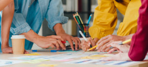 Time de design reunindo ideias sobre produto em uma mesa com diversos papéis coloridos, canetas e lápis para determinar a criação do produto digital. 
