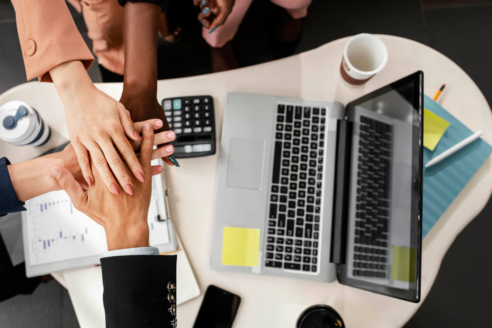 Mãos de profissionais unidas sobre uma mesa de trabalho com laptop e gráficos, representando colaboração estratégica. A imagem ilustra o papel de uma consultoria de experiência do usuário na geração de resultados concretos para os negócios, promovendo decisões mais eficientes e alinhadas à estratégia.